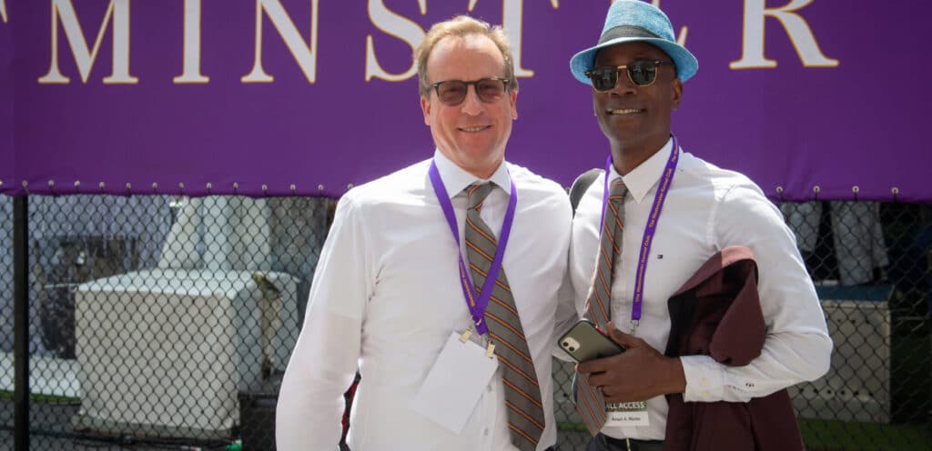 Two men in hats and ties standing in front of a purple sign at the Westminster Kennel Club.