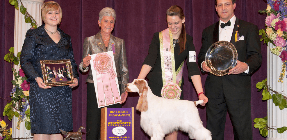 Four people and a dog at an award ceremony with the dog winning a "best junior showmanship" prize.