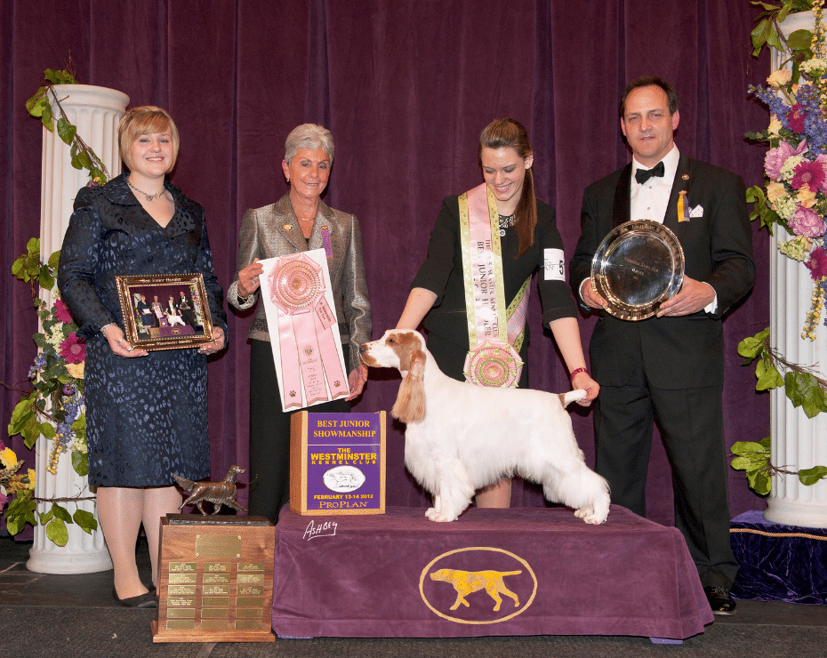 Award recipients at a dog show standing with a white dog and their prizes.