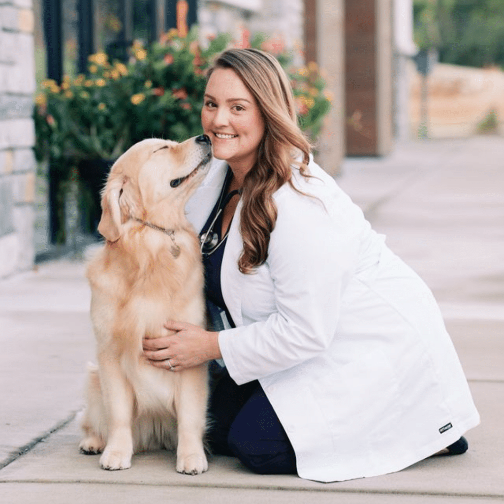 A "Veterinarian of the Year" in a white coat kneels beside a golden retriever on a sidewalk, smiling as the dog licks her face.