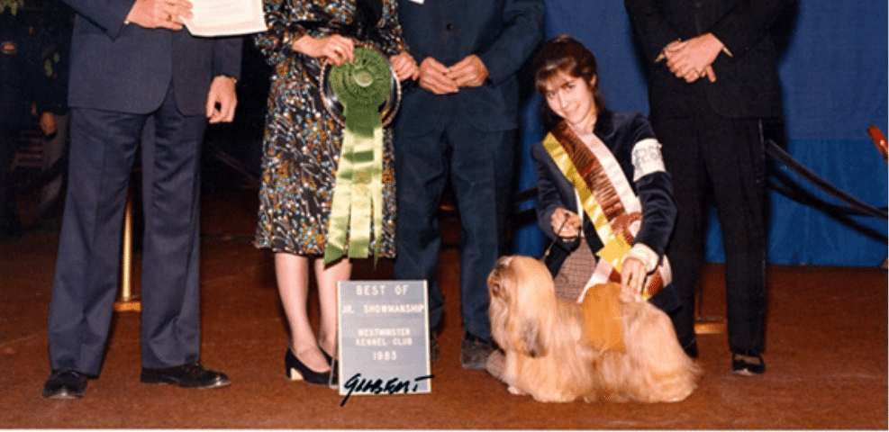 A woman kneels next to a winning dog at a show, holding a trophy and ribbon, with officials standing nearby.