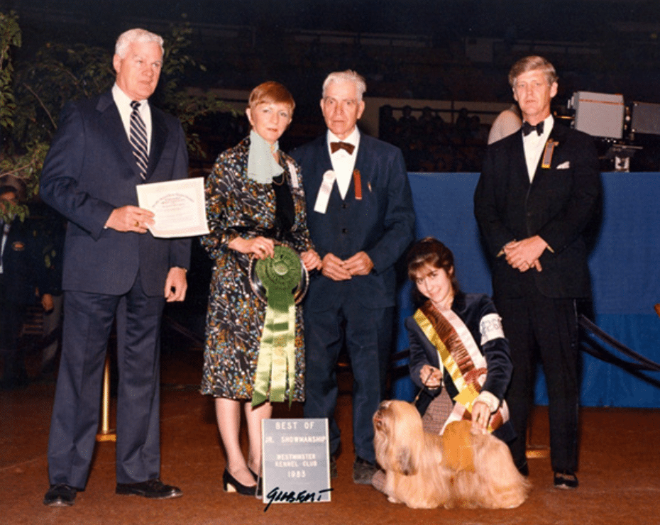 Group of individuals at a dog show with a winner holding a trophy and a ribbon, accompanied by an award-winning dog.