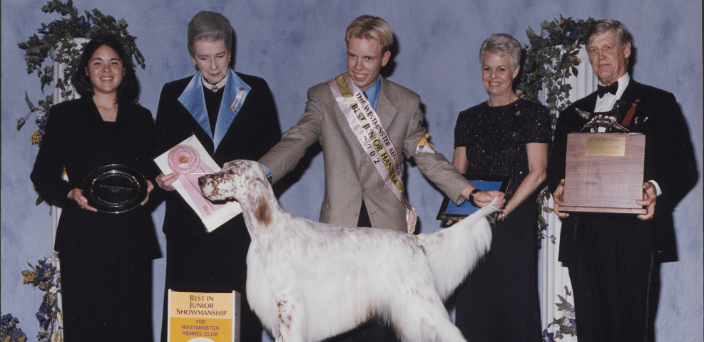 Award recipients and a show dog at a dog show event, with trophies and ribbons on display.