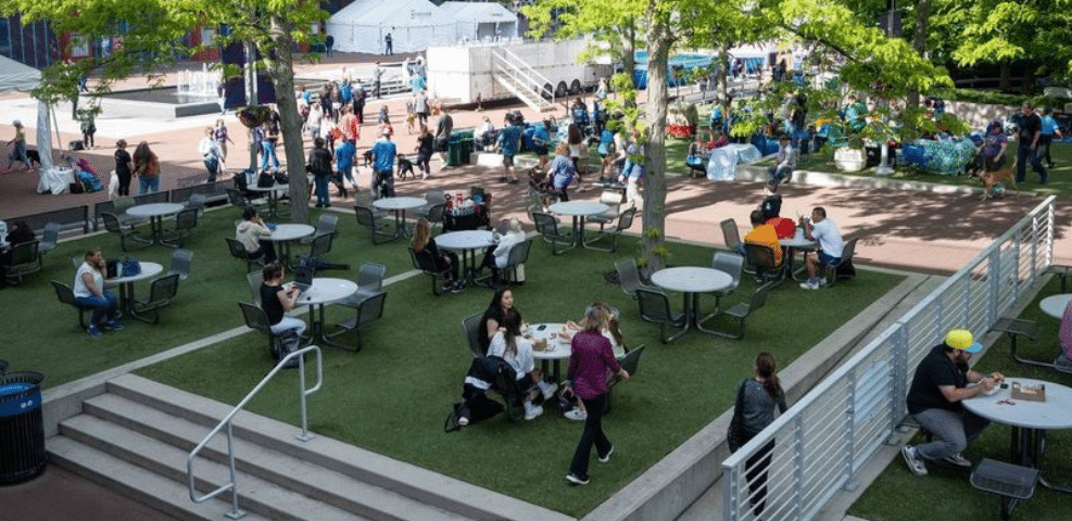 A busy outdoor plaza with exhibitors and people sitting at tables, some conversing and others working on laptops, surrounded by trees and greenery on a sunny day.