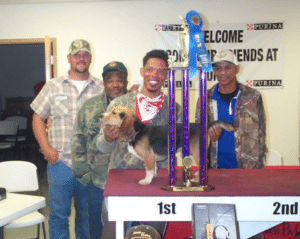 Four men stand behind a table displaying a large trophy and a beagle, with a "Purina" sign in the background. One man, dressed in camouflage and holding the dog, seems to be remembering Kaz Hosaka as he joins the proud presenters.