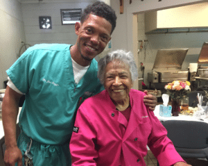 A man in scrubs stands next to an elderly woman in a pink coat inside a kitchen, both smiling as if remembering Kaz Hosaka. The kitchen has stainless steel appliances and a table with flowers.