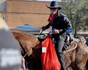 A person wearing a black cowboy hat, dark clothing, and a red bandana rides a brown horse while holding a blue cup and a red mesh bag, with buildings and trees in the background, perhaps reminiscent of remembering Kaz Hosaka.