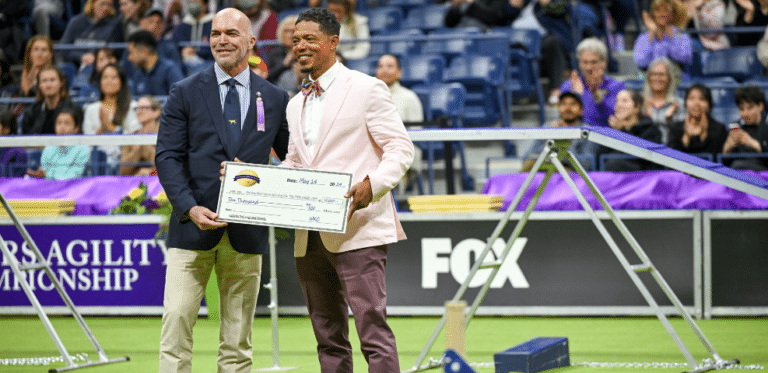 Two men in formal attire holding a large check stand in an indoor dog agility competition arena with an audience in the background, perfectly capturing a moment of celebration for our blog.
