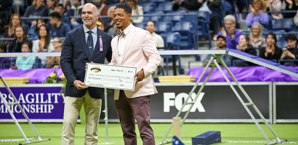 Two men in formal attire holding a large check stand in an indoor dog agility competition arena with an audience in the background, perfectly capturing a moment of celebration for our blog.