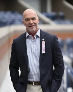 A bald man in a striped shirt and blazer stands outdoors with hands in pockets, wearing a ribbon on his lapel. The background features a blurred stadium setting.
