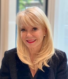 A person with long blonde hair and a black top smiles while sitting indoors in front of a window.