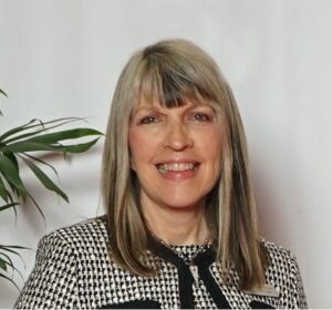 Smiling woman with long hair in a houndstooth pattern jacket stands near a leafy plant against a white background.