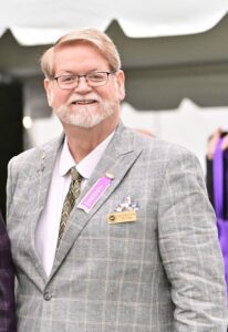 A man with glasses, a beard, and a mustache is wearing a gray checkered suit with a patterned tie and name tag, standing outdoors.