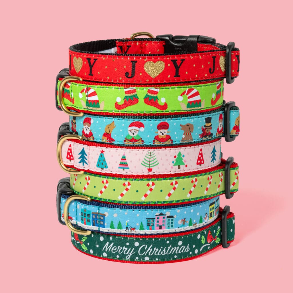 A stack of six festive dog collars with Christmas-themed designs on a pink background.