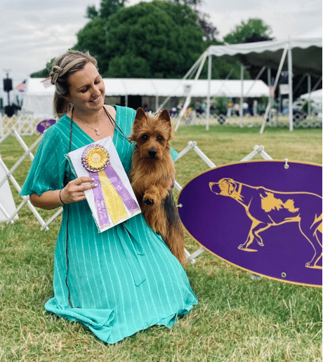 A woman kneels on grass holding a small brown dog and a ribbon award. A purple sign with a dog silhouette is nearby. Tents and trees are in the background.