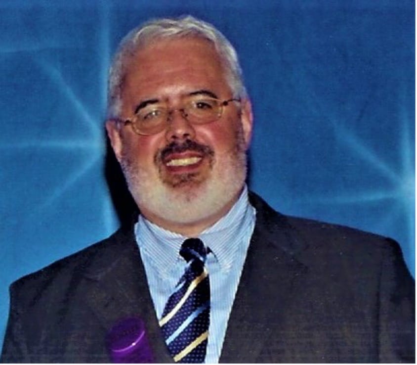A smiling man with glasses and a white beard holds a purple object while standing against a blue background, wearing a suit and striped tie.
