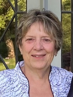 A smiling person with short hair stands in front of a window, wearing a white top with a dark pattern. Trees are visible in the background through the window.