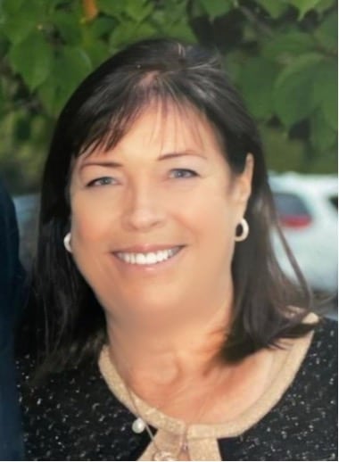 Smiling woman with dark hair wearing a black and beige top, standing outdoors with greenery in the background.