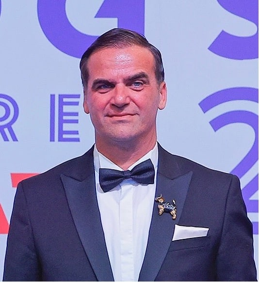 A person in a tuxedo with a bow tie stands in front of a backdrop featuring letters and numbers.