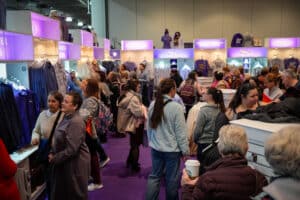 People browsing apparel in a crowded merchandise booth with purple displays at an indoor event, mingled with your guide to the 149th Annual Westminster Kennel Club Dog Show.