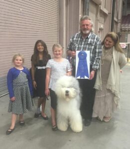 A group of people stand with a fluffy white dog, holding a large blue ribbon.