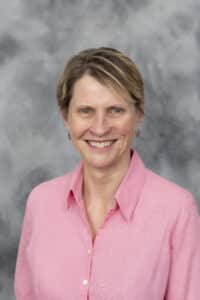 A woman with short blonde hair wearing a light pink collared shirt smiles in front of a gray mottled background.