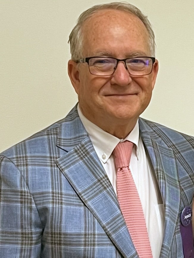 An older man, possibly one of the judges, wearing glasses, a blue plaid suit jacket, a white shirt, and a light pink tie stands in front of a plain light-colored wall.