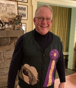 A smiling man wearing glasses and a dark vest with a purple and yellow ribbon design stands indoors near a stone fireplace and framed pictures, appearing ready to impress the judges.