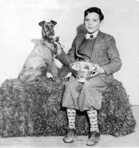 A boy in a suit sits on a hay bale holding a metal bowl, with a dog beside him. The dog has a ribbon on its collar and one paw on the boy’s arm.