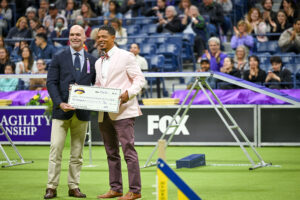 Two men in suits stand on a turf field holding a large ceremonial check, with an audience seated in bleachers behind them at an agility championship event focused on promoting canine health.