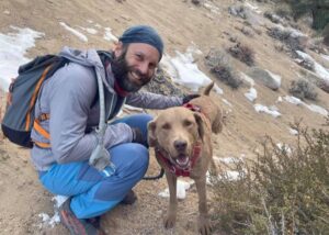 A man in outdoor gear kneels next to a brown dog from a breed rescue on a sandy, snowy trail, with rocks and shrubs in the background.