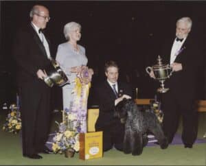 Four judges in formal attire stand beside a handler kneeling with a winning dog and large ribbon at a dog show; trophies and flowers are displayed nearby.
