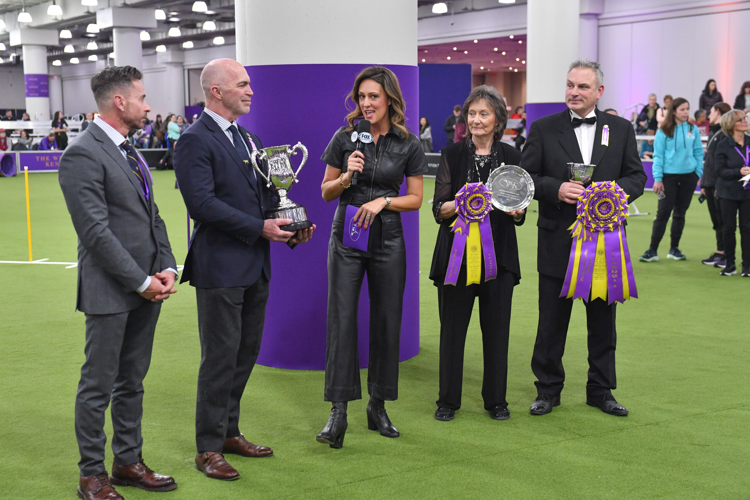 A woman interviews three judges holding trophies and ribbons at an indoor dog show event, as people watch in the background. Meet Dr. Sara Lyle: The Westminster Kennel Club’s 2025 Veterinarian of the Year.