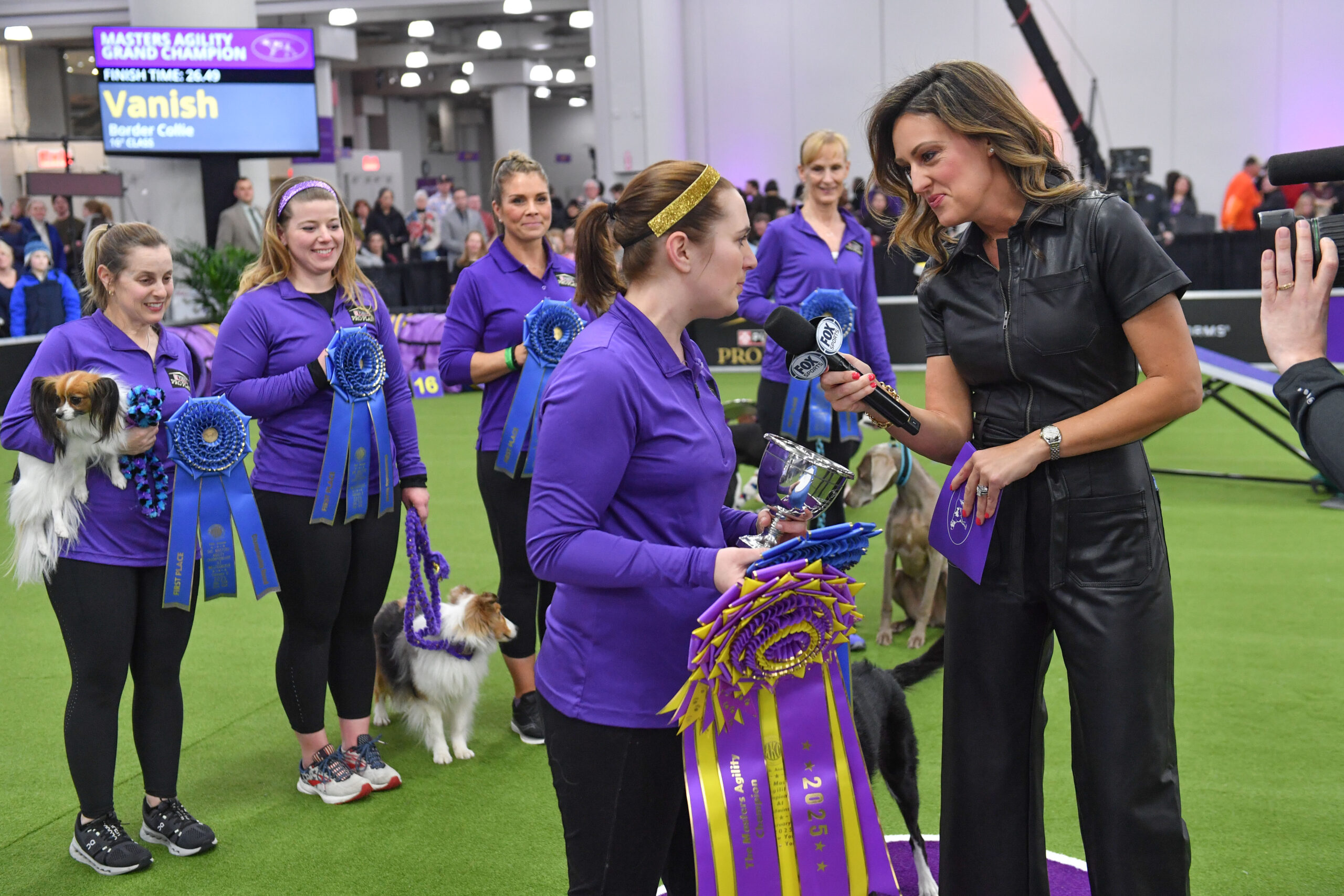A dog agility competition winner receives trophies and ribbons from an interviewer, while other contestants with their dogs stand in the background. Meet Dr. Sara Lyle: The Westminster Kennel Club’s 2025 Veterinarian of the Year.