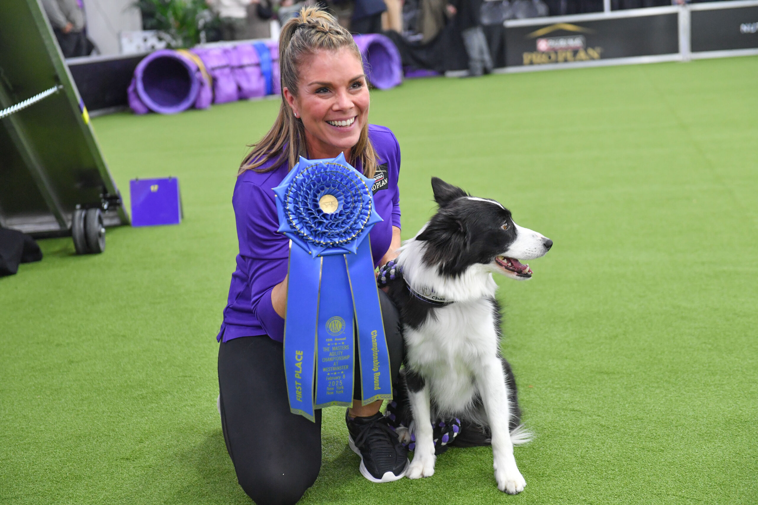 Meet Dr. Sara Lyle: The Westminster Kennel Club’s 2025 Veterinarian of the Year, who kneels on green turf holding a large blue first place ribbon beside a black and white dog at a competition event.