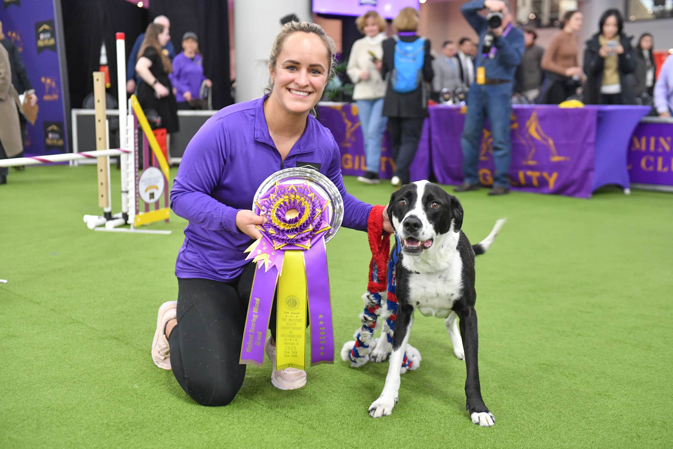 Meet Dr. Sara Lyle: The Westminster Kennel Club’s 2025 Veterinarian of the Year, as she kneels beside a black and white dog, proudly holding a large purple and yellow award ribbon at an indoor dog agility competition.