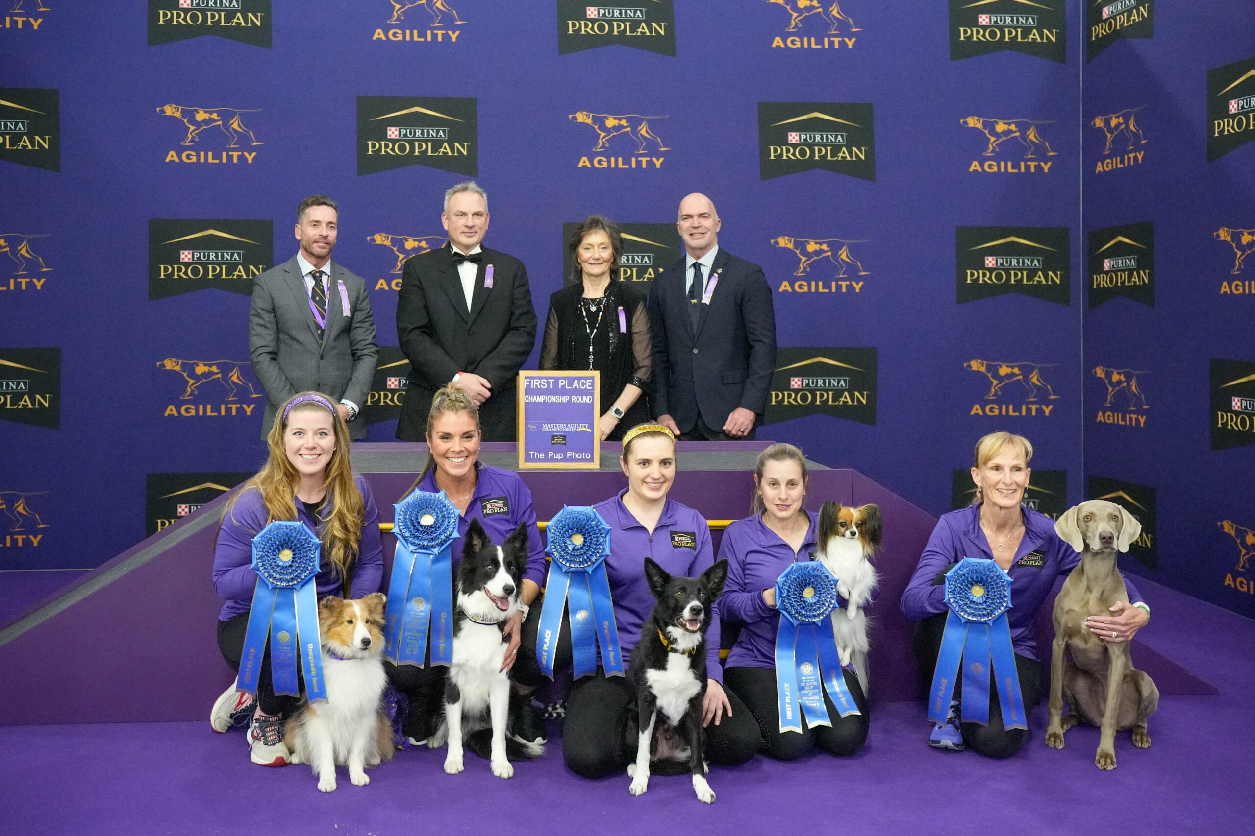 Group of people and their dogs pose with large blue first place ribbons at a Purina Pro Plan Agility event, standing in front of a purple background featuring event branding and celebrating Meet Dr. Sara Lyle: The Westminster Kennel Club’s 2025 Veterinarian of the Year.