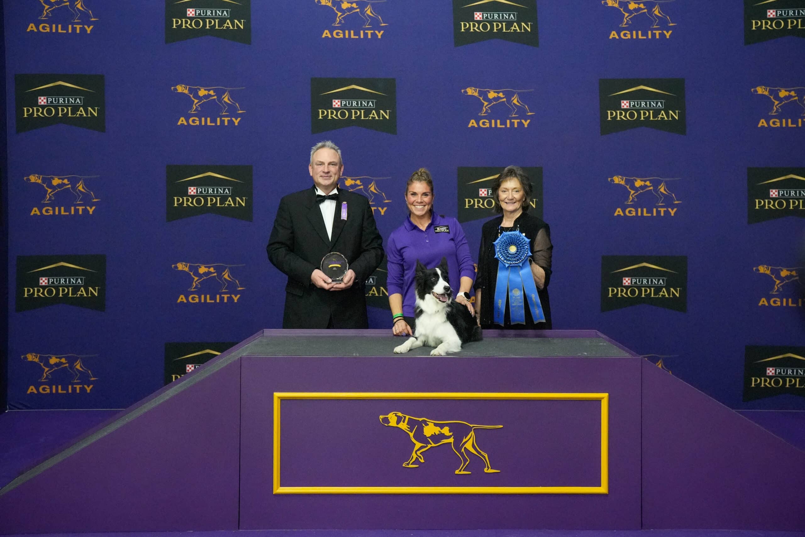 Meet Dr. Sara Lyle: The Westminster Kennel Club’s 2025 Veterinarian of the Year, as three people and a black-and-white dog pose on a platform at a dog agility competition, with awards and branded signage in the background.