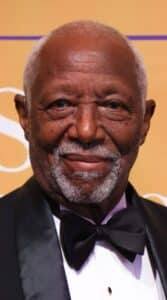 An older man with gray hair and a beard wearing a black tuxedo and bow tie, smiling in front of a gold background.
