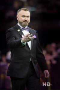 A man in a black tuxedo with a purple bow tie and pocket square gestures with his hand while standing indoors, capturing the elegance of History and Traditions: Looking Back as we Look Forward to 150.
