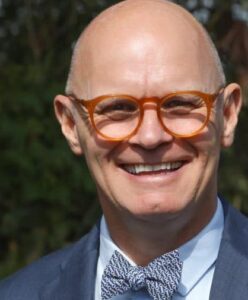 A bald man wearing round orange glasses, a blue suit, light blue shirt, and patterned bow tie smiles outdoors.