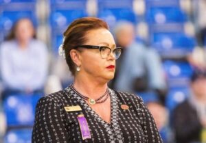 Woman with glasses and red lipstick wearing a black and white patterned outfit, standing indoors with blue seats blurred in the background.