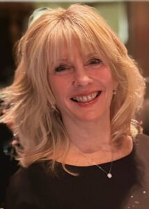 A woman with shoulder-length blonde hair and bangs smiles at the camera, wearing a black top and a necklace with a small pendant.