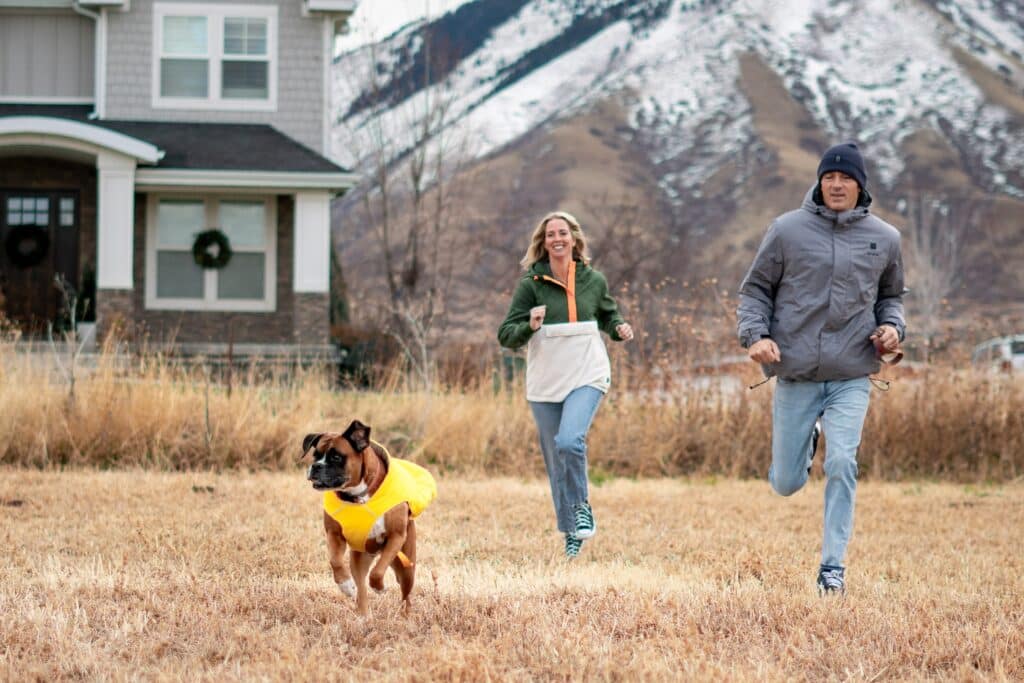 A man, woman, and dog in a yellow vest run across a grassy field in front of a house, with snow-covered mountains in the background, as they discuss the 2026 Vendor Application Form.