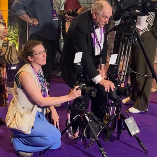 Two photographers, one kneeling and one standing, adjust their cameras on tripods at an event with a purple carpet and other people in the background.