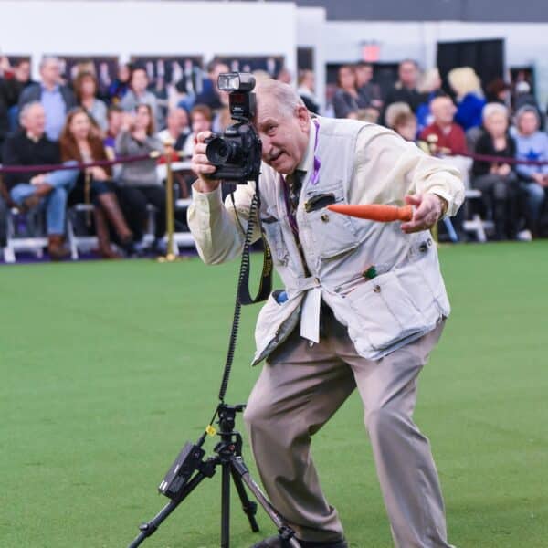 A photographer in a white vest kneels on green turf, holding a camera and a dog toy, with a tripod nearby and an audience seated in the background.