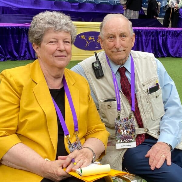 An older woman in a yellow blazer and an older man in a vest with credentials sit together at an indoor event with a purple backdrop.