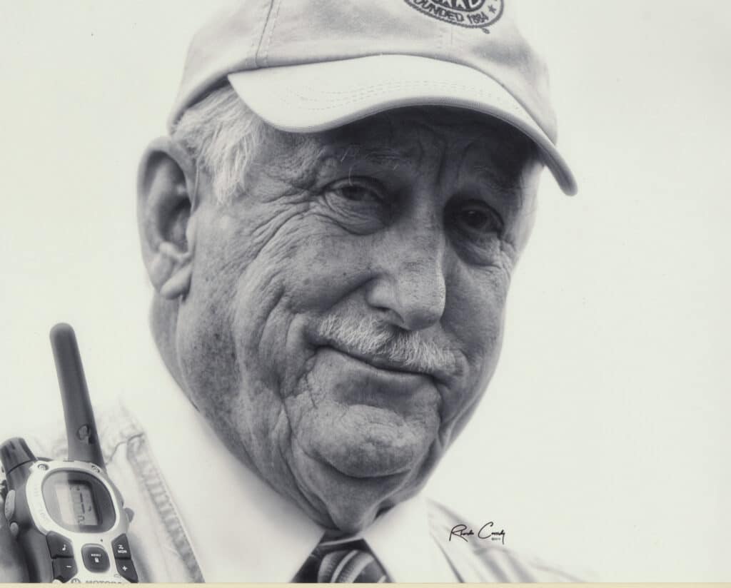 An older man wearing a cap and collared shirt looks at the camera, holding a walkie-talkie. The image is in black and white.