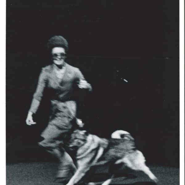 A person in a suit walks a dog on a leash across a dark stage or floor. The image is blurred, suggesting motion.