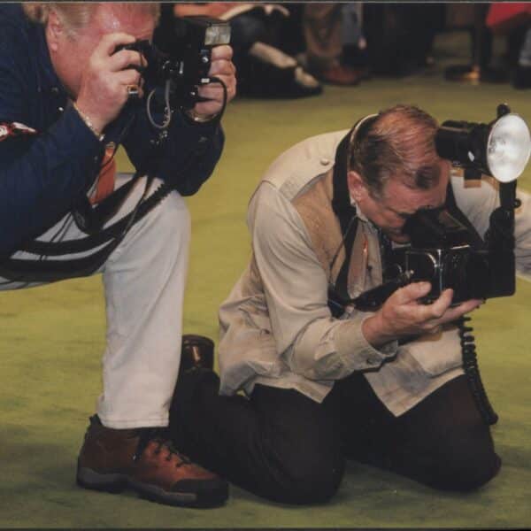 Two photographers crouch on a green carpet, each holding cameras with large flashes, focused intently as they take photos.